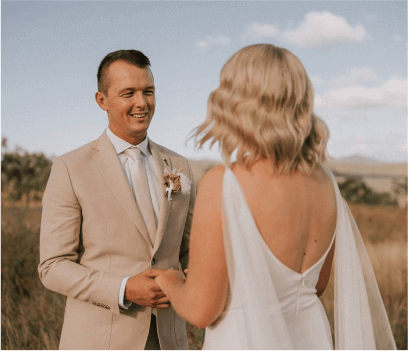 Pareja de novios en una ceremonia al aire libre, sonriendo y mirándose a los ojos, rodeados de un paisaje natural con un cielo despejado. El novio lleva un traje claro y la novia un vestido blanco con un velo.