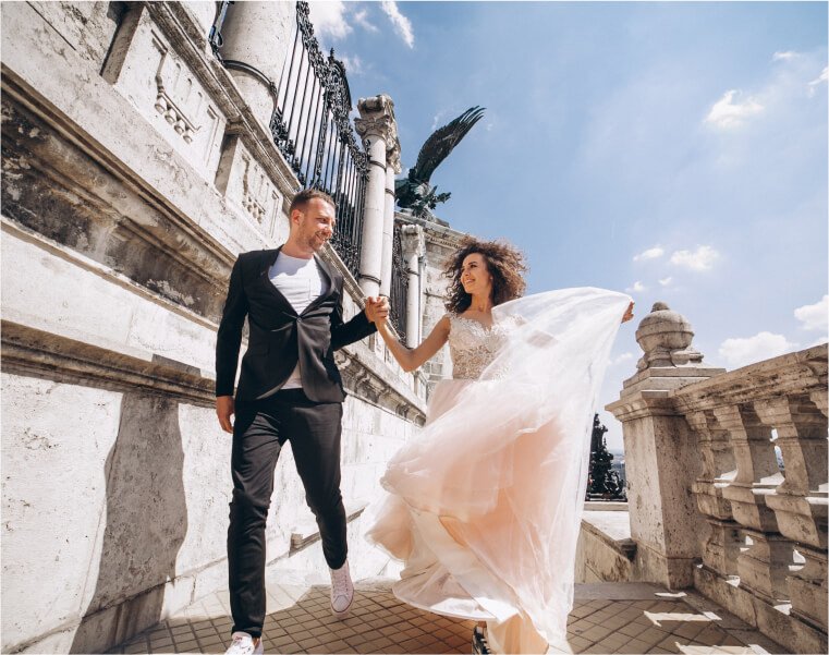 Pareja de recién casados caminando de la mano en un entorno arquitectónico, con el vestido de la novia ondeando al viento y una expresión de felicidad.