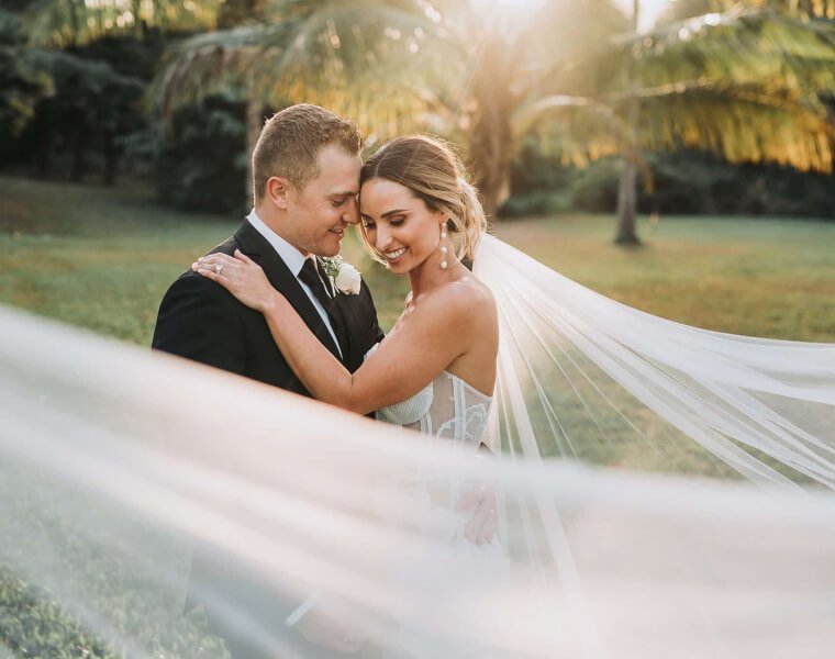 Pareja de novios abrazándose en un hermoso entorno al aire libre, con luz suave del atardecer y un velo de novia ondeando.