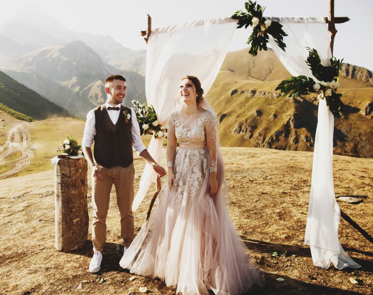 Pareja de novios sonrientes en una ceremonia de boda al aire libre, con un fondo montañoso y un arco decorado con telas y vegetación. La novia viste un vestido largo y elegante, mientras que el novio lleva un atuendo casual con chaleco.