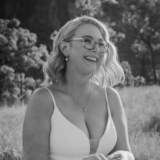 Mujer sonriendo en un campo, con un vestido blanco y gafas, rodeada de naturaleza.
