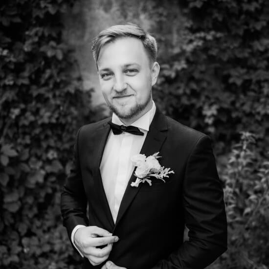 Hombre vestido con traje de gala y pajarita, posando frente a un fondo de hojas verdes, con una flor en la solapa. La imagen es en blanco y negro, destacando la elegancia y el estilo del sujeto.