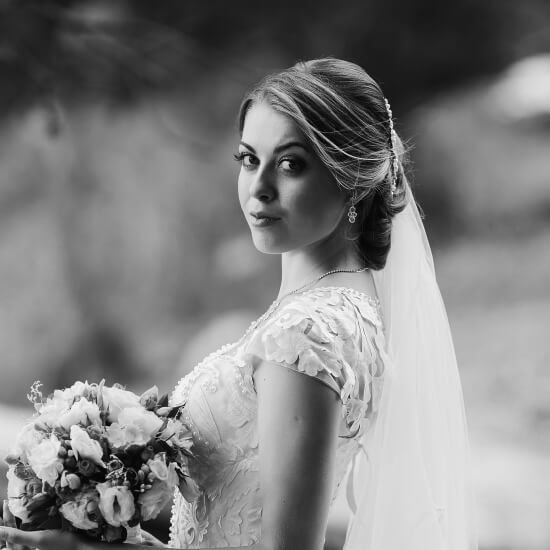 Novia en vestido blanco con encaje, sosteniendo un ramo de flores, posando en un entorno natural. La imagen es en blanco y negro, destacando su expresión serena y el velo.