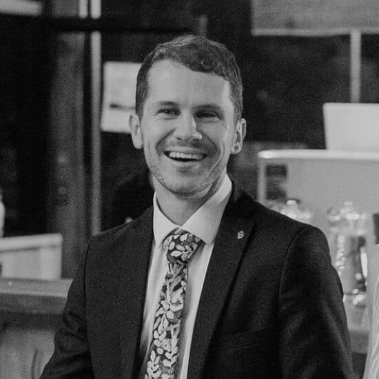 Hombre sonriendo con traje oscuro y corbata estampada, sentado en un ambiente elegante.