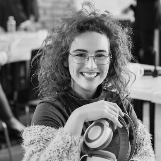 Mujer sonriente con cabello rizado y gafas, sosteniendo unos auriculares, en un ambiente acogedor y moderno.