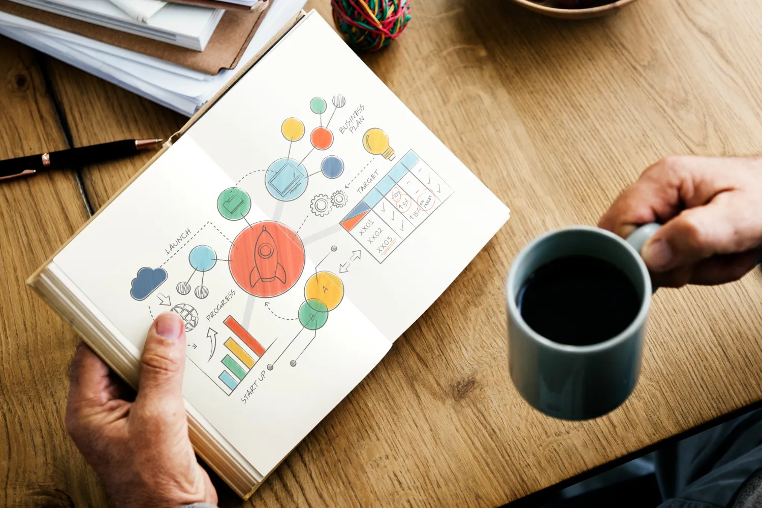 Mano sosteniendo una taza de café mientras examina un cuaderno abierto con diagramas coloridos sobre planificación empresarial, incluyendo un cohete, gráficos de progreso y objetivos. Fondo de mesa de madera con documentos apilados.